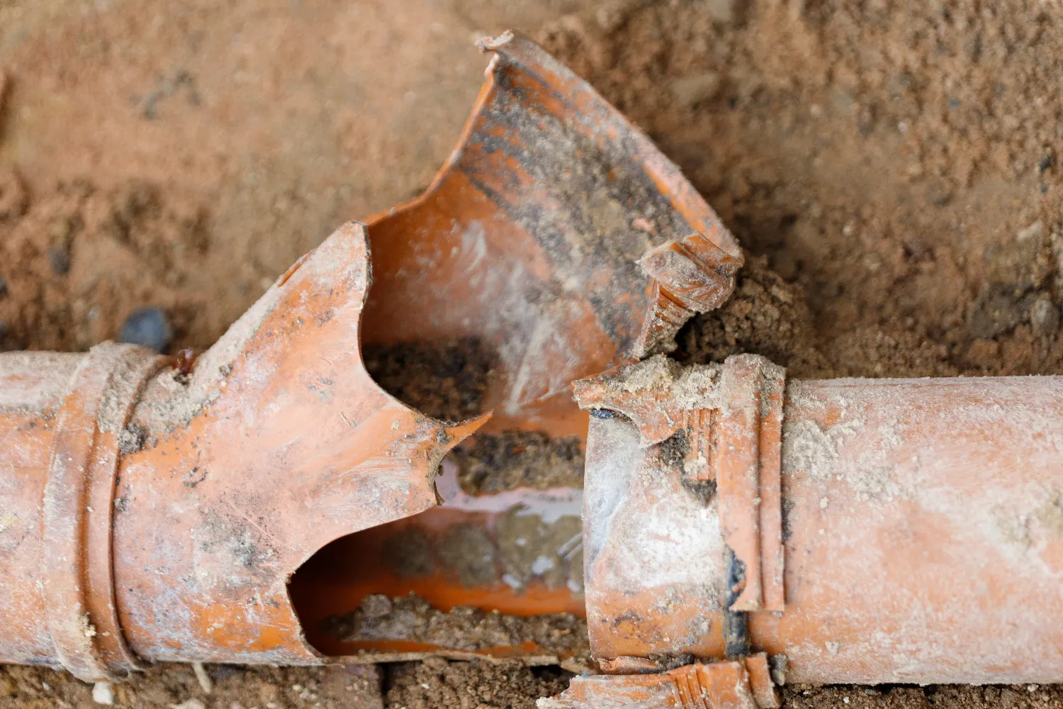 A close-up of a broken orange clay pipe surrounded by soil, showing significant cracks and damage with fragments missing.