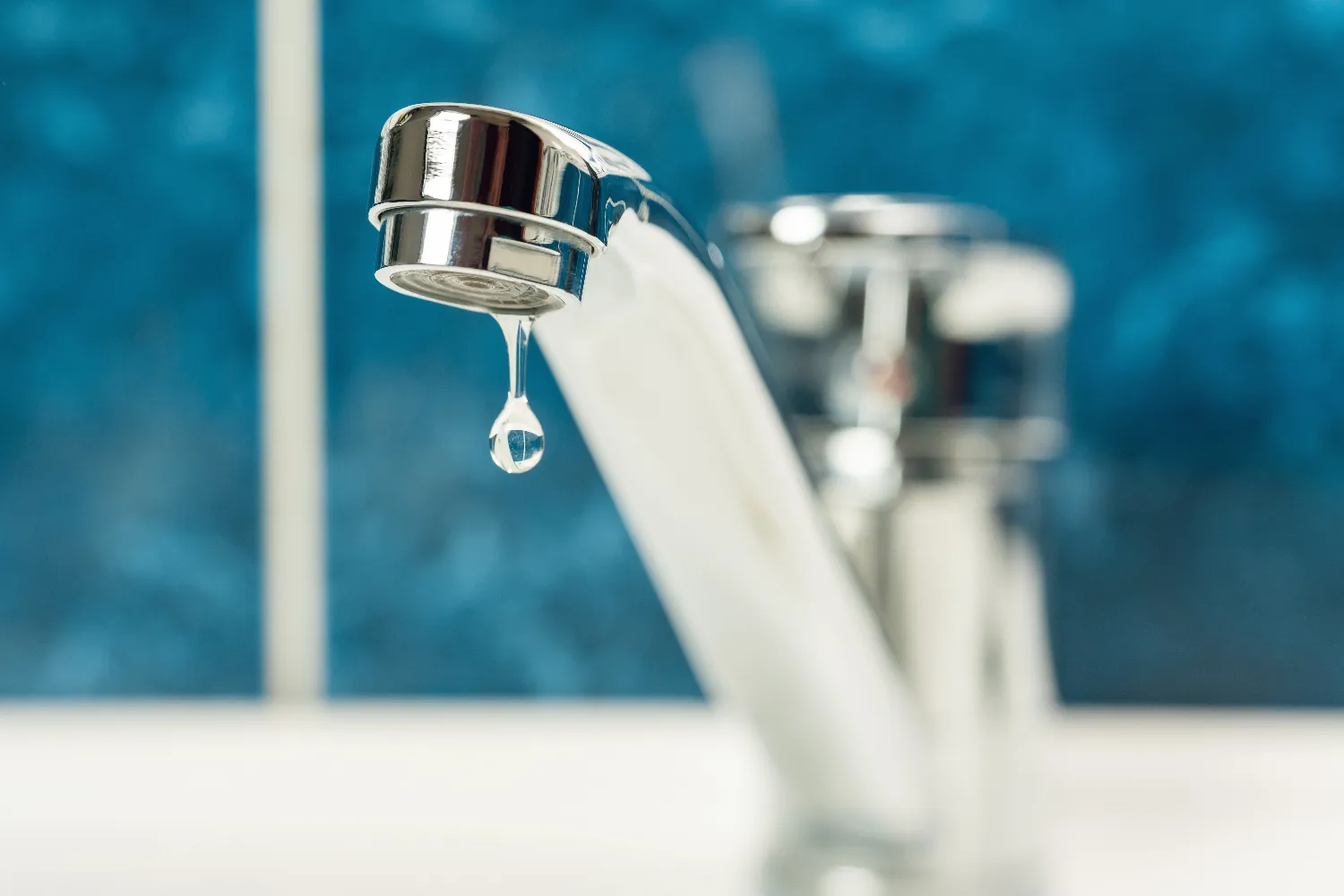 A close-up of a chrome faucet with a single water droplet hanging from the spout, set against a blue and white tiled background—a perfect reminder of the importance of timely faucet leak repair.