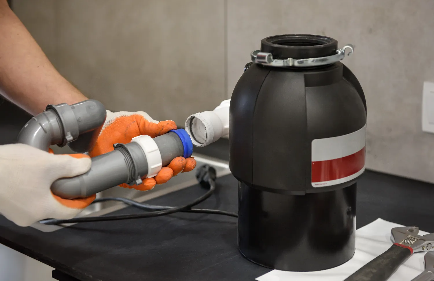 Person wearing gloves is connecting grey PVC pipes to a black garbage disposal unit on a kitchen countertop. A wrench and power cable are visible nearby, highlighting the process of installing or repairing a garbage disposal system.