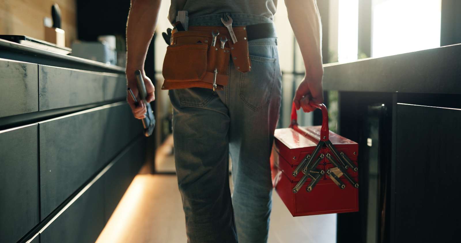 A person wearing a tool belt and jeans holds a red toolbox in one hand and a tool in the other, walking through a modern kitchen, ready to tackle any kitchen plumbing task.
