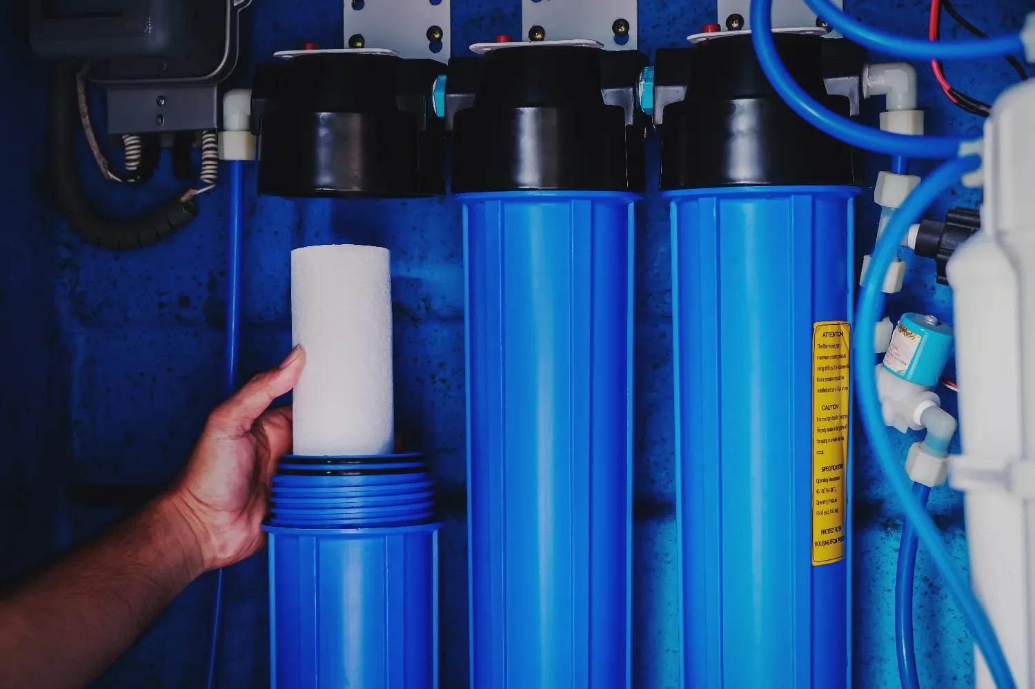 A person inserts a white filter cartridge into a blue water filtration system, with two identical blue filter housings beside it, emphasizing the process of clean water purification.
