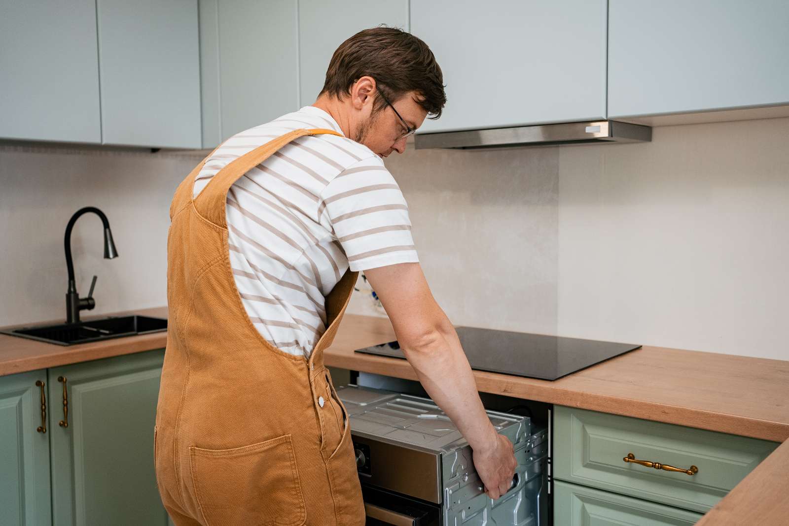 A person in overalls installs or removes a dishwasher in a modern kitchen with light green cabinets, a black sink, and attends to kitchen plumbing.