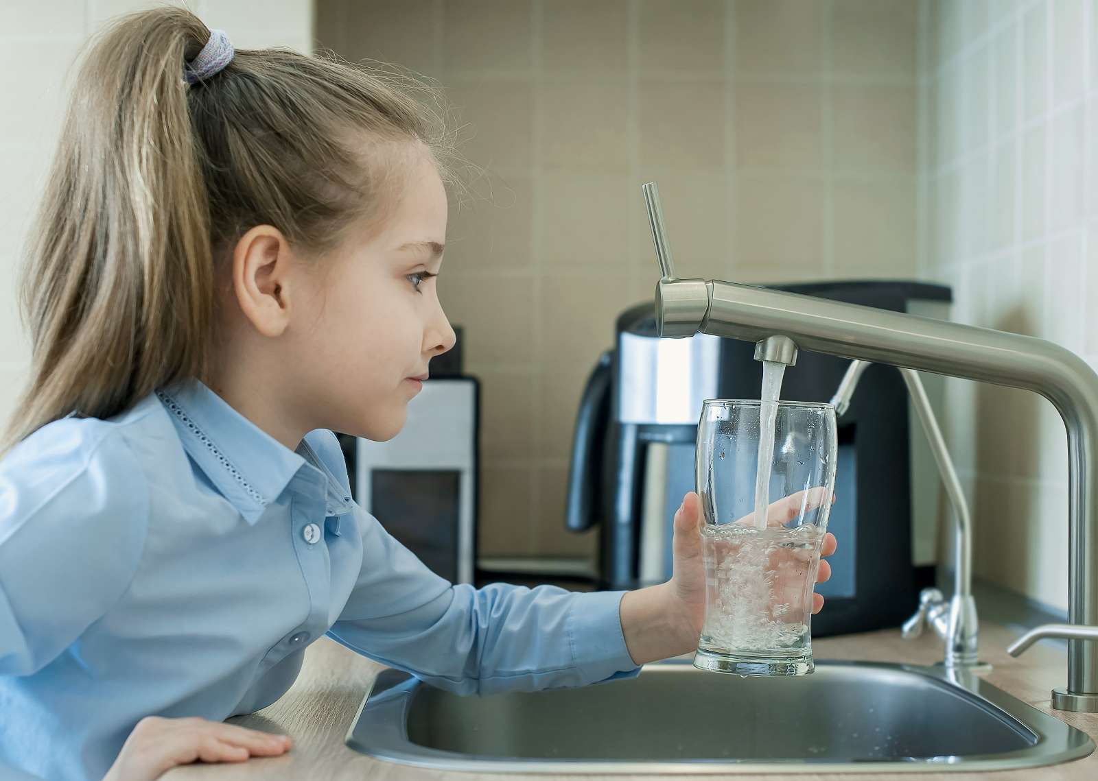 A young girl fills a glass with water from a kitchen faucet, standing at the sink in a modern kitchen equipped with water purification.